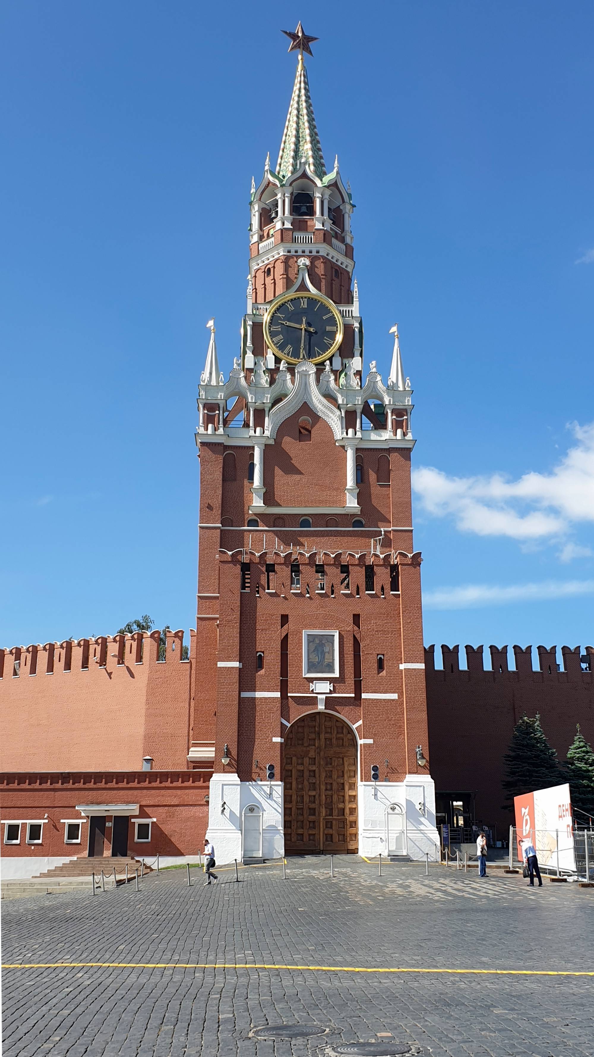 Красная кирпичная башня с часами и звездой на шпиле, часть Кремлёвской стены, под ясным голубым небом; у входа — несколько человек, справа — рекламный щит.