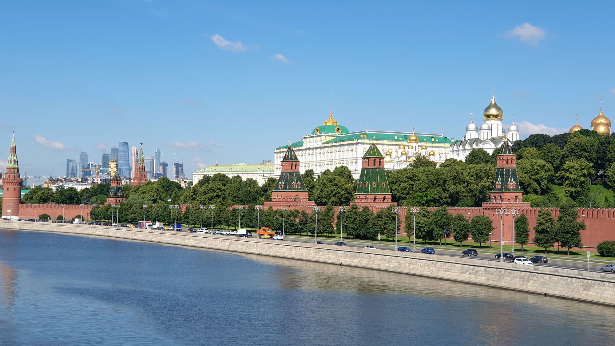 Вид на Московский Кремль с набережной: красные стены и башни, Большой Кремлёвский дворец с золотыми куполами, соборы и современные небоскрёбы на фоне голубого неба.