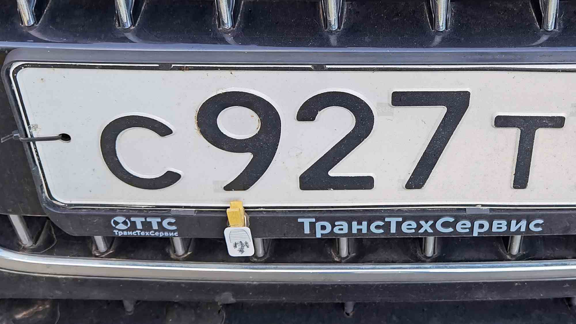 Автомобильный номер С927ТТ в рамке с надписью «ТрансТехСервис» и логотипом «ТТС», закреплённый на решётке радиатора автомобиля.