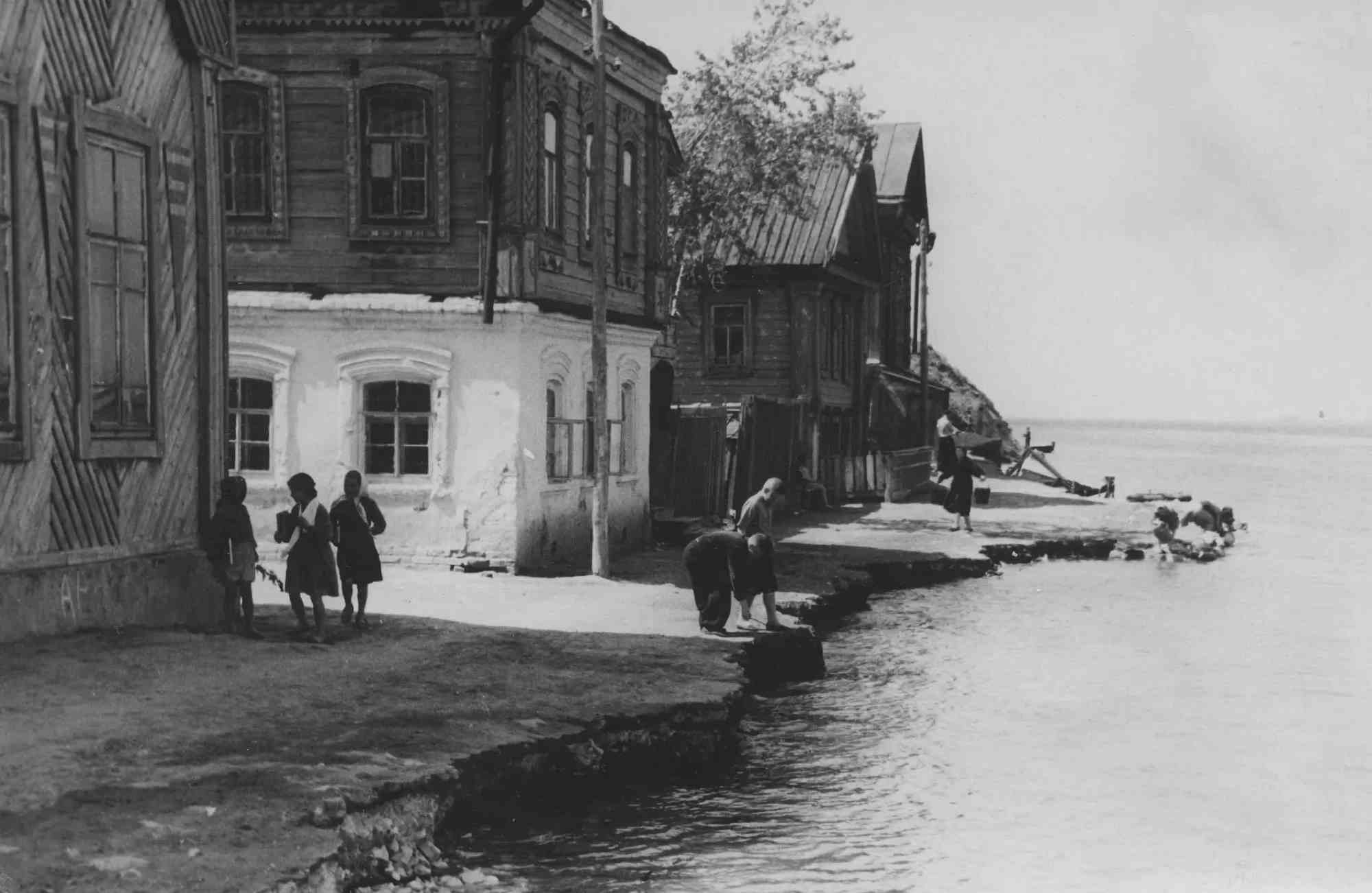 Чёрно-белое фото: у берега реки стоят деревянные дома, дети и взрослые моют бельё у воды, на заднем плане — лодка и склон.