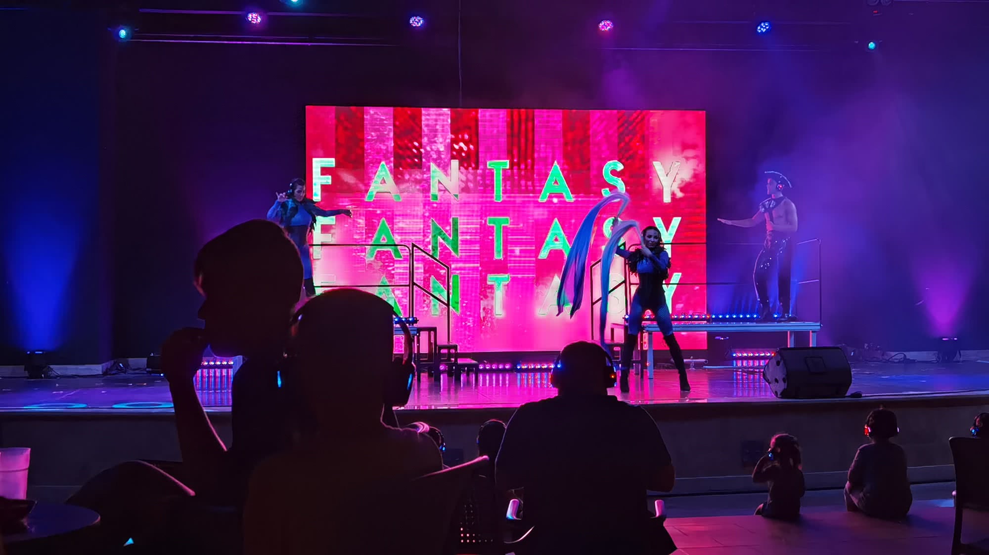 Танцоры в костюмах выступают на сцене с неоновым экраном «FANTASY» на фоне, зрители в наушниках наблюдают за шоу в полумраке.