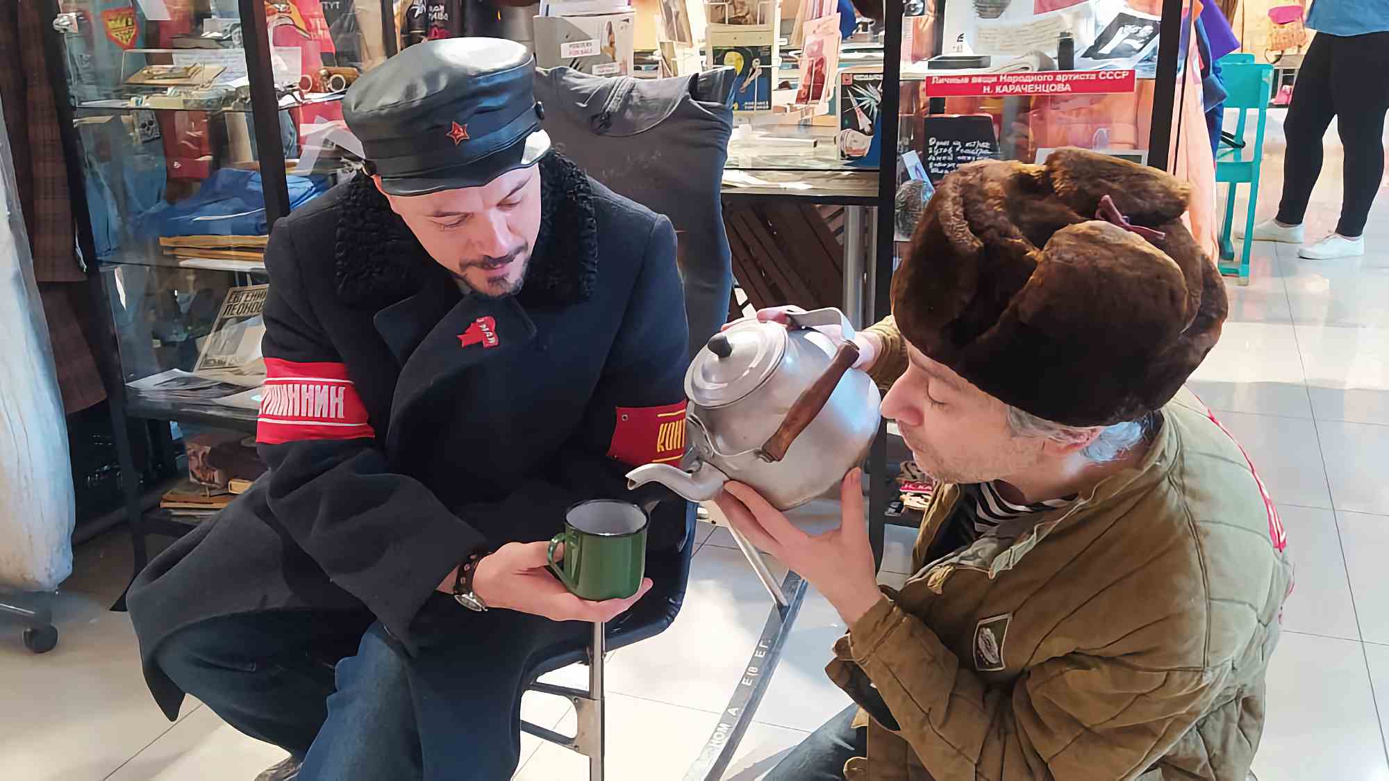 Два человека в советской военной форме: один в фуражке с красной звездой и нарукавной повязкой «Комиссар» наливает чай из чайника в кружку, другой в ушанке наблюдает.