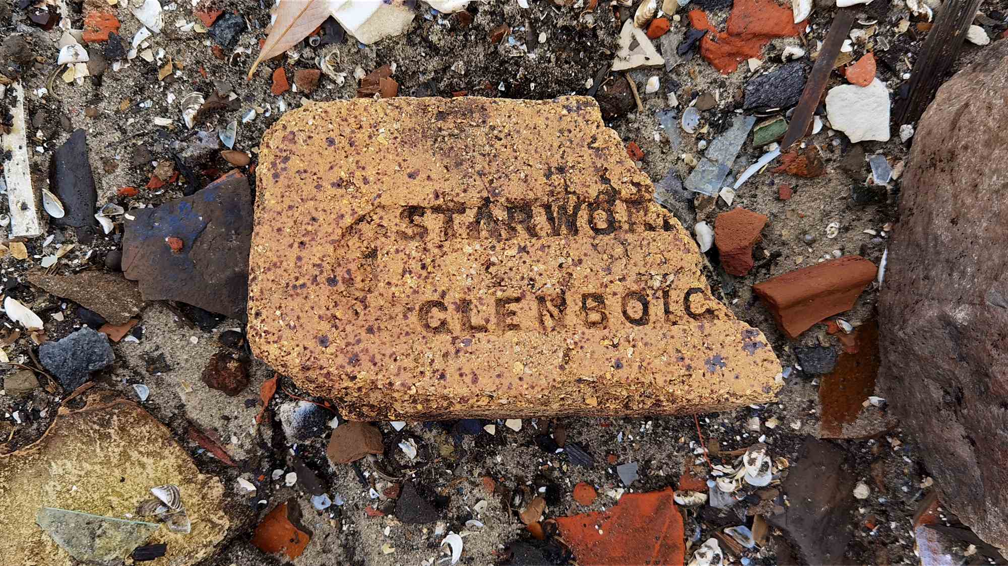 Обломок кирпича с выгравированными словами «STARWORTH» и «GLENBOIG», лежащий среди осколков керамики, стекла и ракушек на земле.