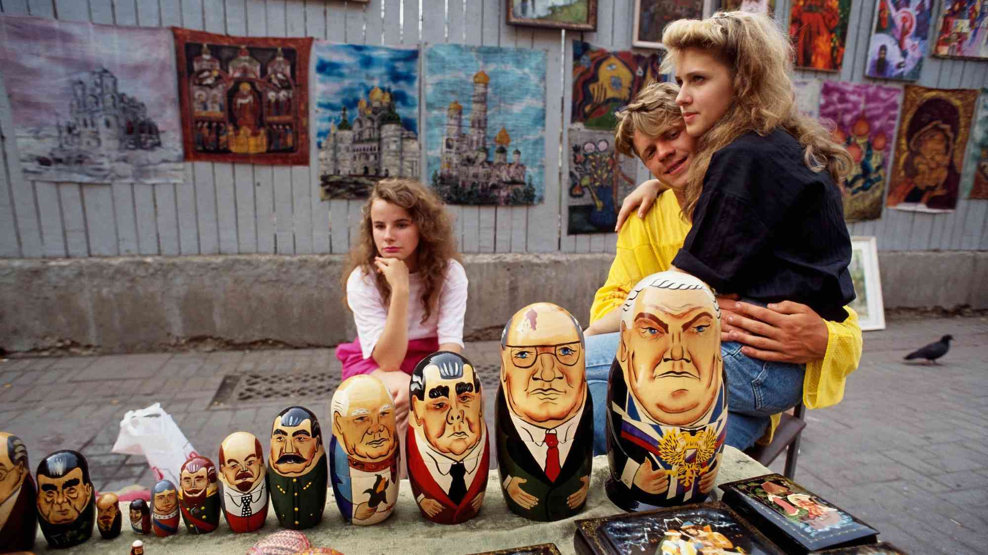 Три молодых человека сидят за столом с матрёшками, расписанными под портреты российских политиков, на фоне вывесок с картинами храмов и икон.