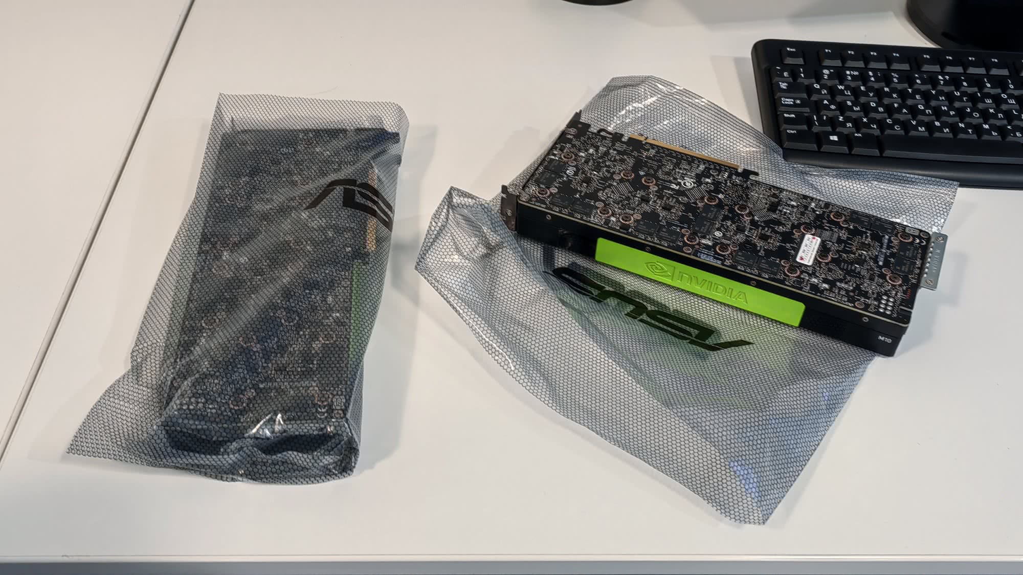Видеокарта NVIDIA с зелёной полоской и логотипом ASUS в антистатическом пакете на белом столе рядом с клавиатурой.