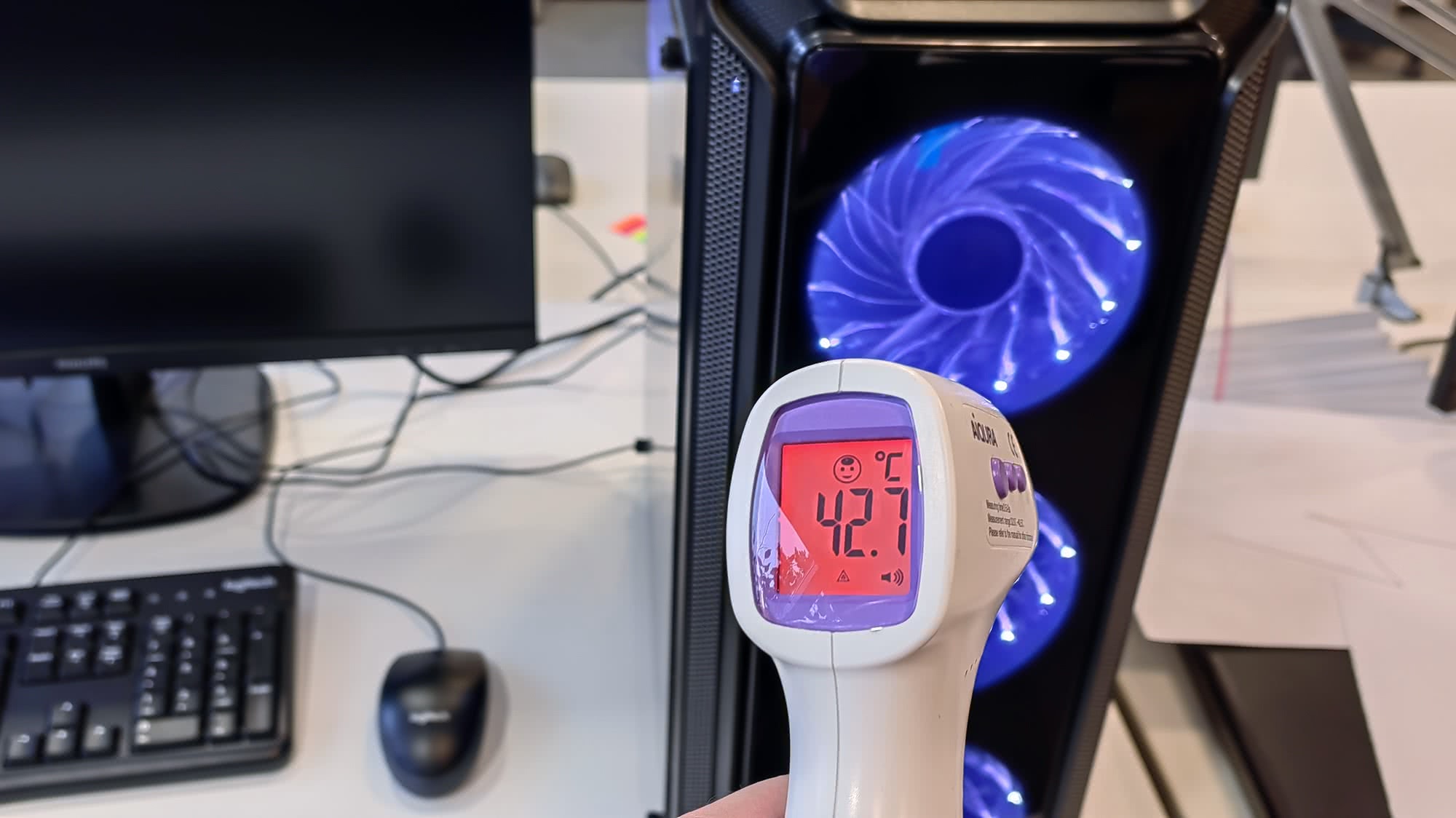 Бесконтактный термометр показывает температуру 42,7 °C при измерении корпуса компьютерного системного блока с синей подсветкой вентиляторов.