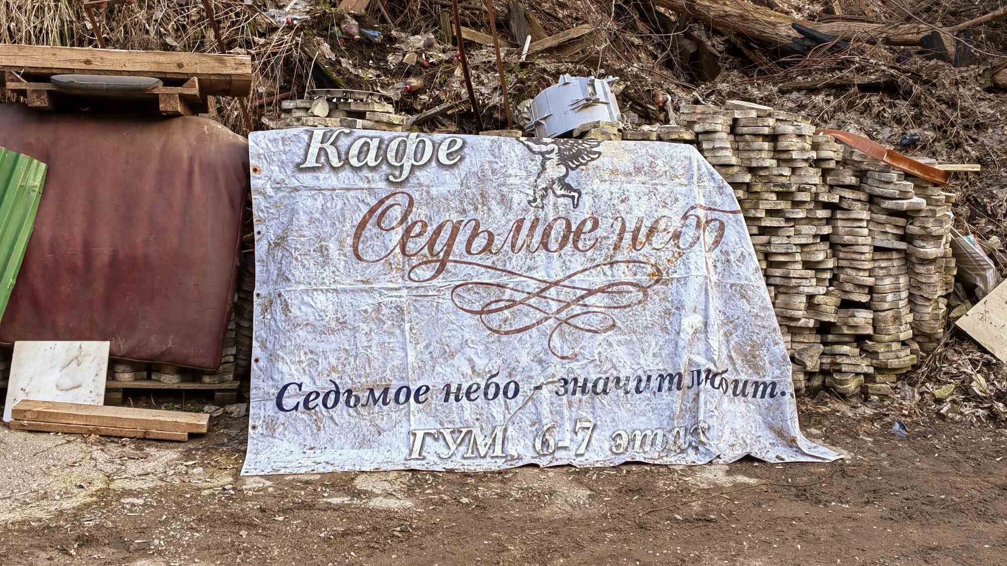 Грязный вывеска кафе «Седьмое небо» с надписью «Седьмое небо — значит любит. ГУМ, 6-7 этаж», лежащая на земле среди мусора и строительных отходов.