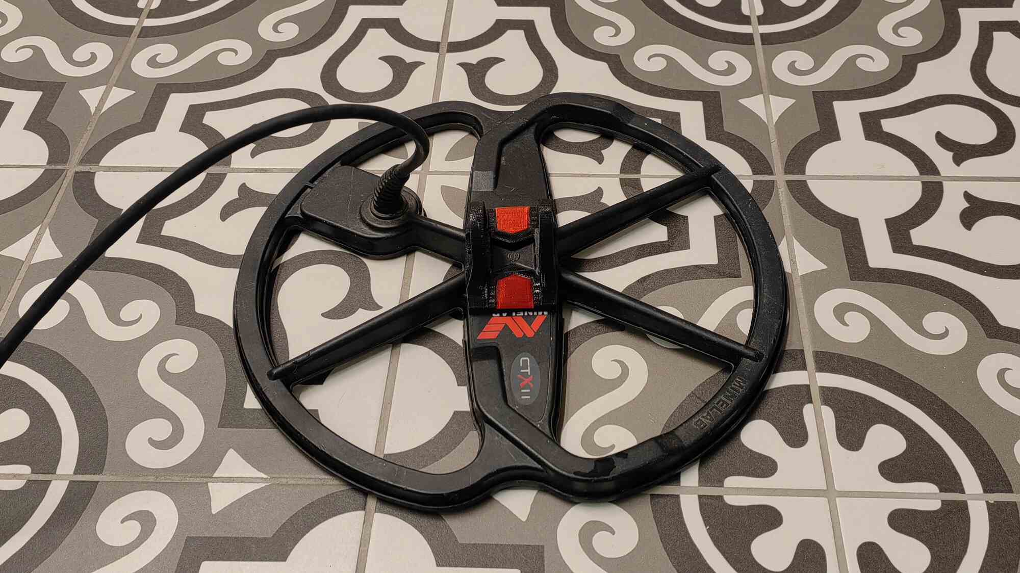 Чёрная круглая катушка металлоискателя Minelab CTX II с красными элементами и кабелем, лежащая на плитке с серо-белым орнаментом.