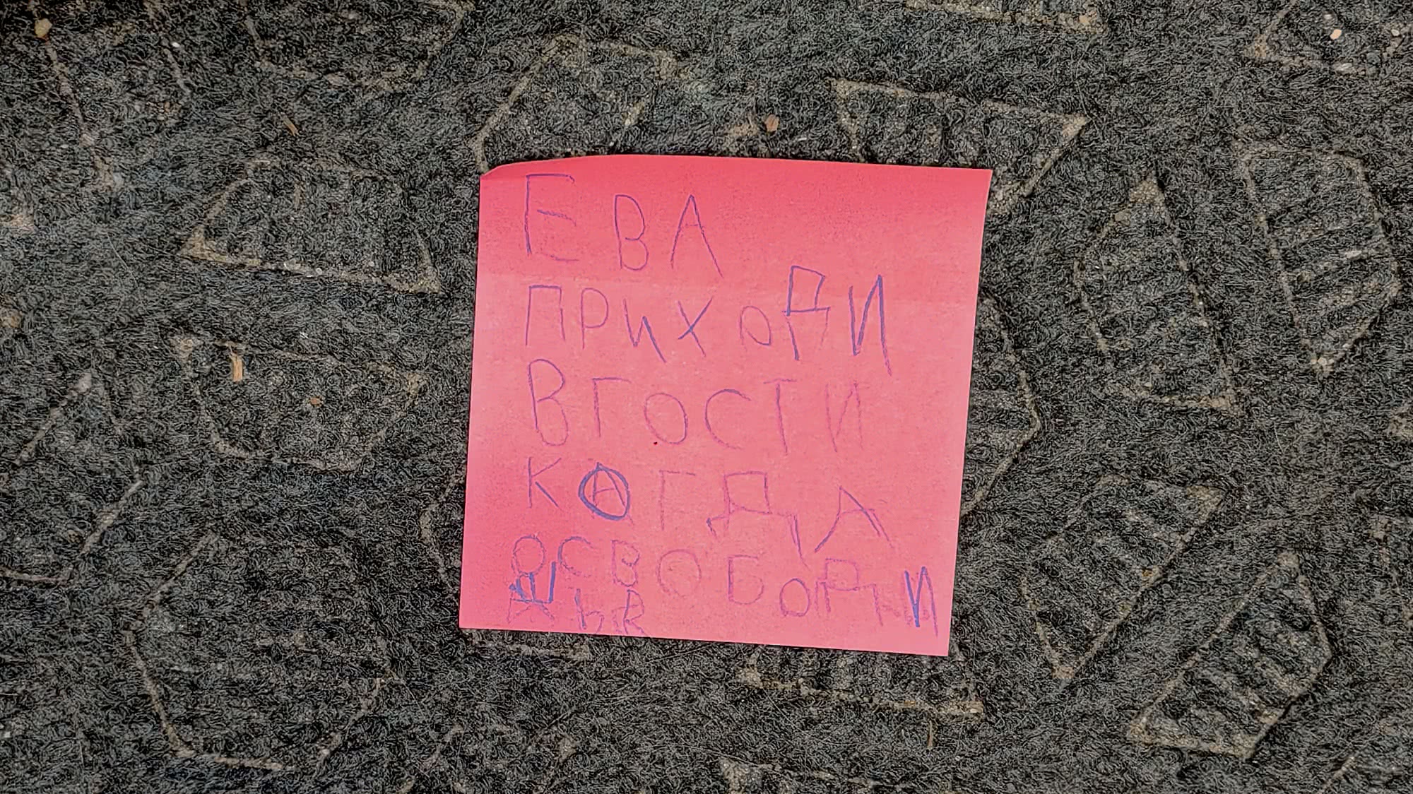 Розовая записка с рукописным текстом: «ЕВА ПРИХОДИ В ГОСТИ КОГДА ОСВОБОДИШЬСЯ», лежащая на тёмной фактурной поверхности.