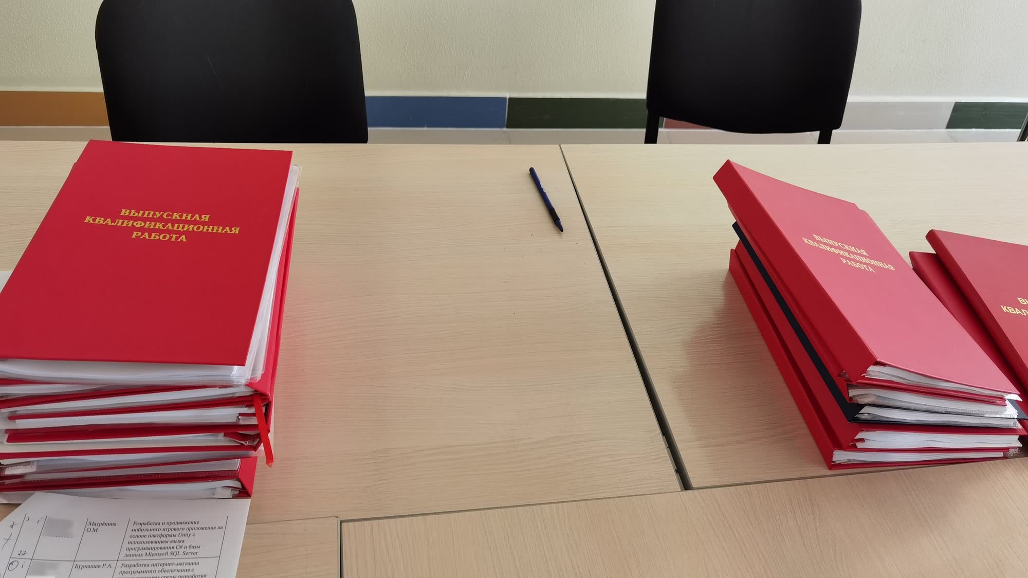 Стопки красных папок с надписью «ВЫПУСКНАЯ КВАЛИФИКАЦИОННАЯ РАБОТА» на светлом столе, рядом ручка и лист с таблицей; на заднем плане — два чёрных стула.