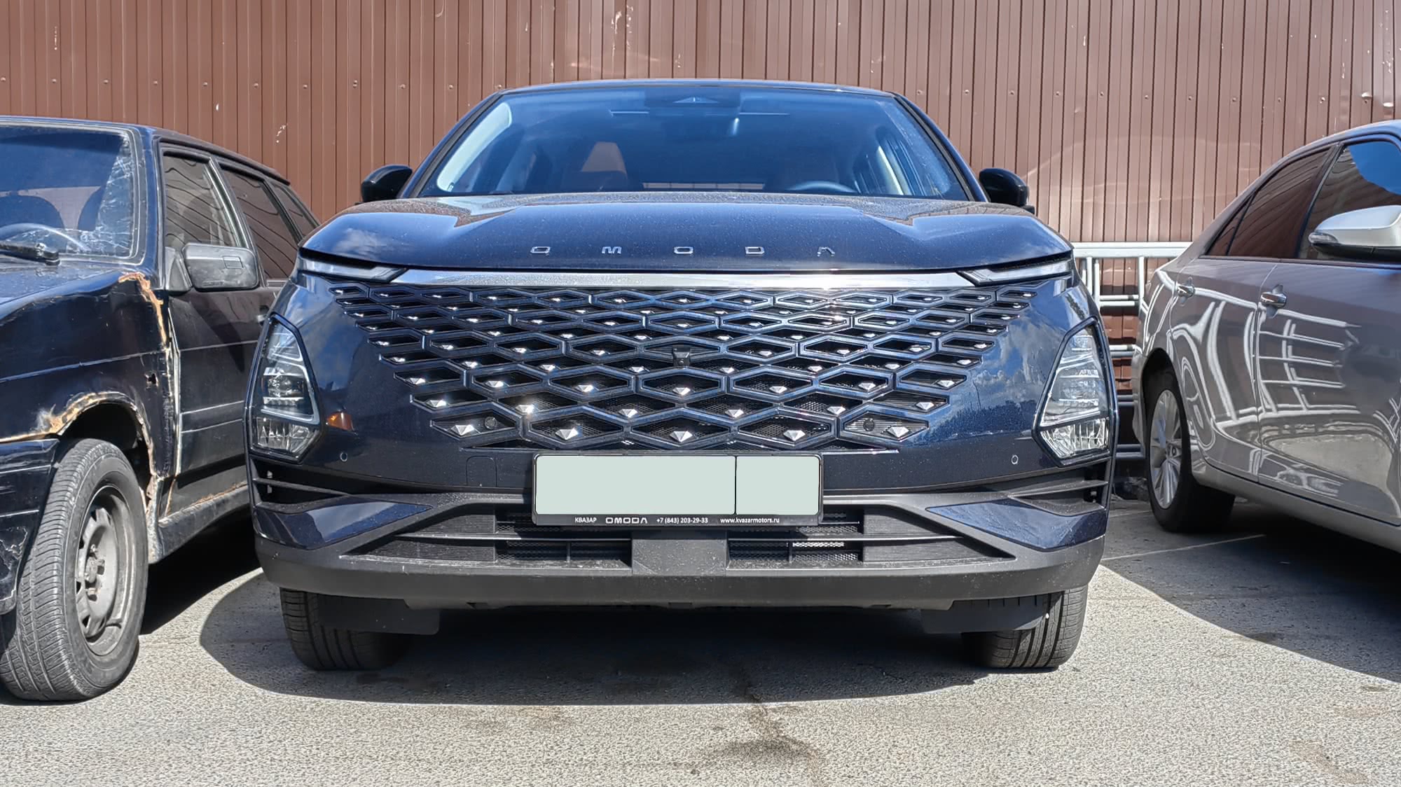 Чёрный кроссовер OMODA с крупной шестиугольной решёткой радиатора и надписью бренда на капоте, припаркованный на асфальте между двумя другими автомобилями у коричневой стены.