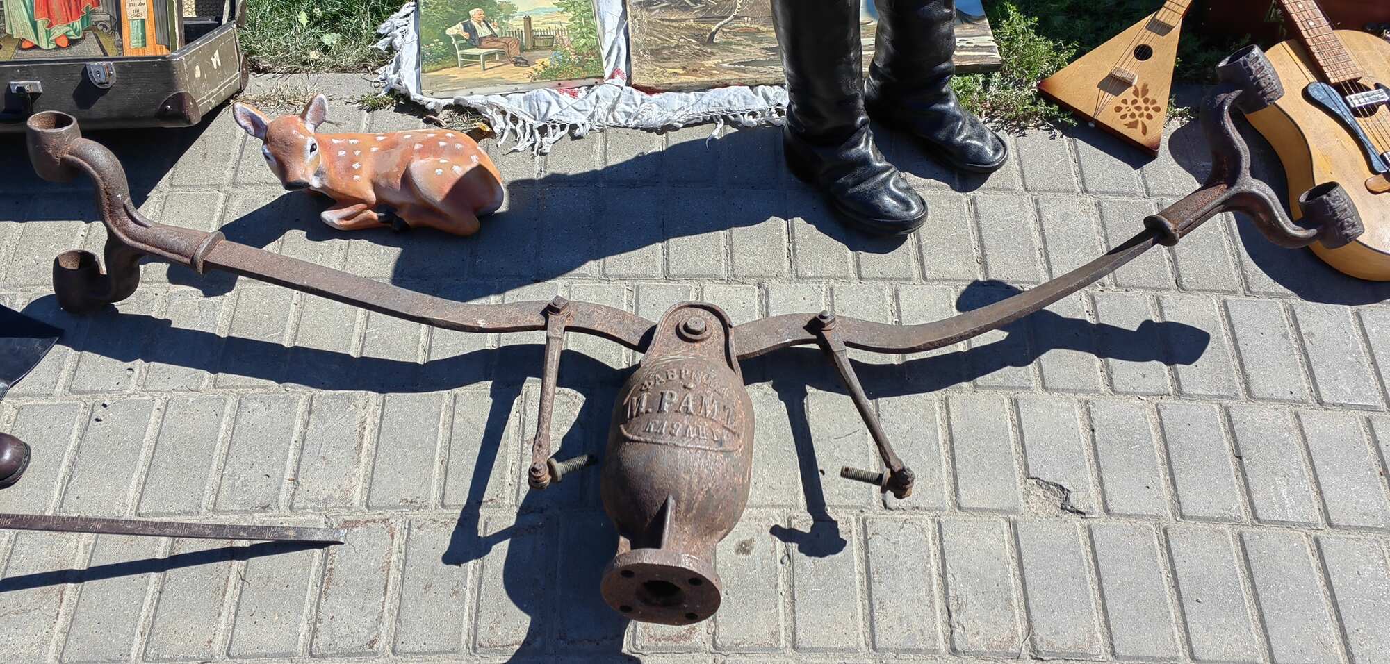 Ржавый металлический механизм с надписью «М. РАММ» на корпусе лежит на брусчатке; рядом — фигурка оленя, ботинки, балалайка и гитара.