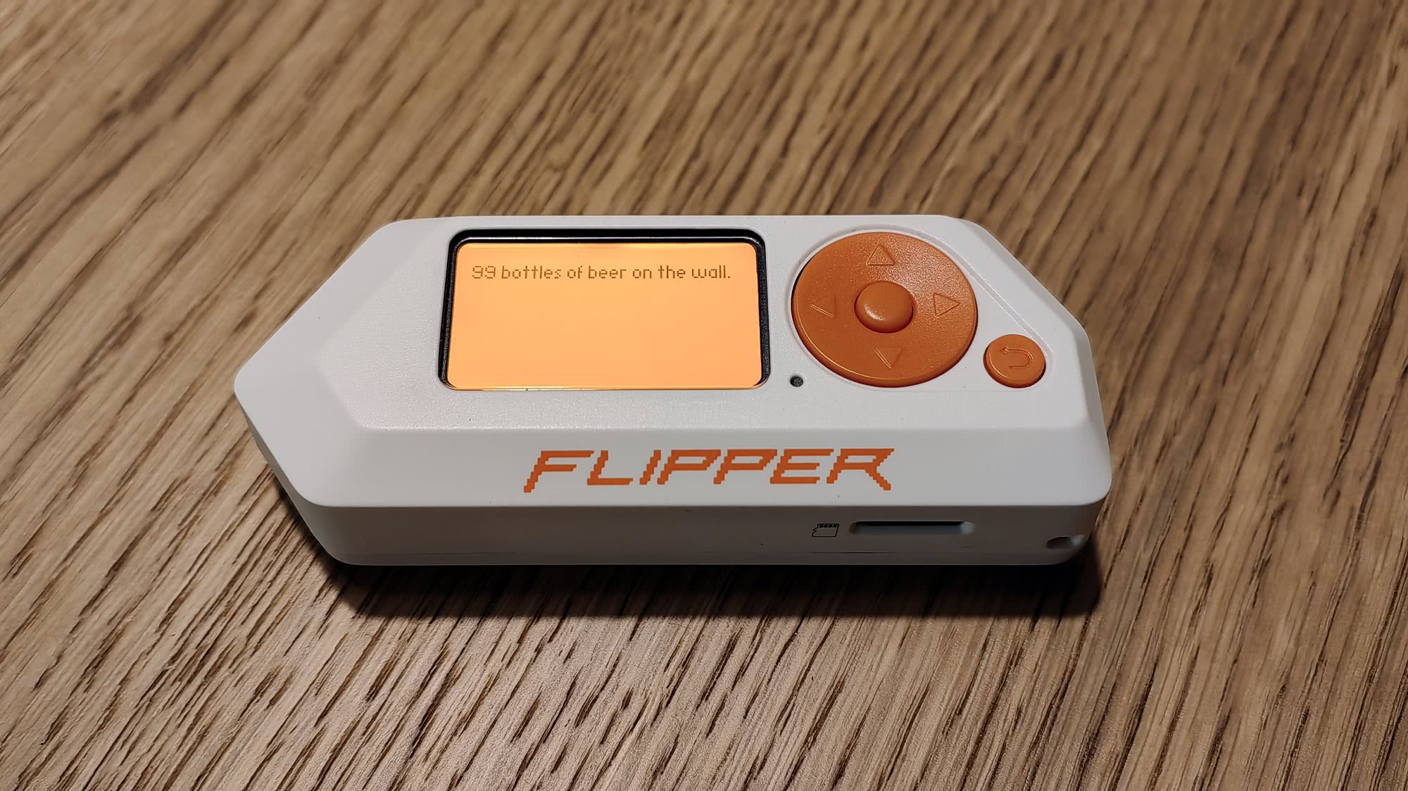 Белый многофункциональный инструмент Flipper Zero с оранжевым экраном, на котором написано «99 bottles of beer on the wall», лежит на деревянной поверхности.