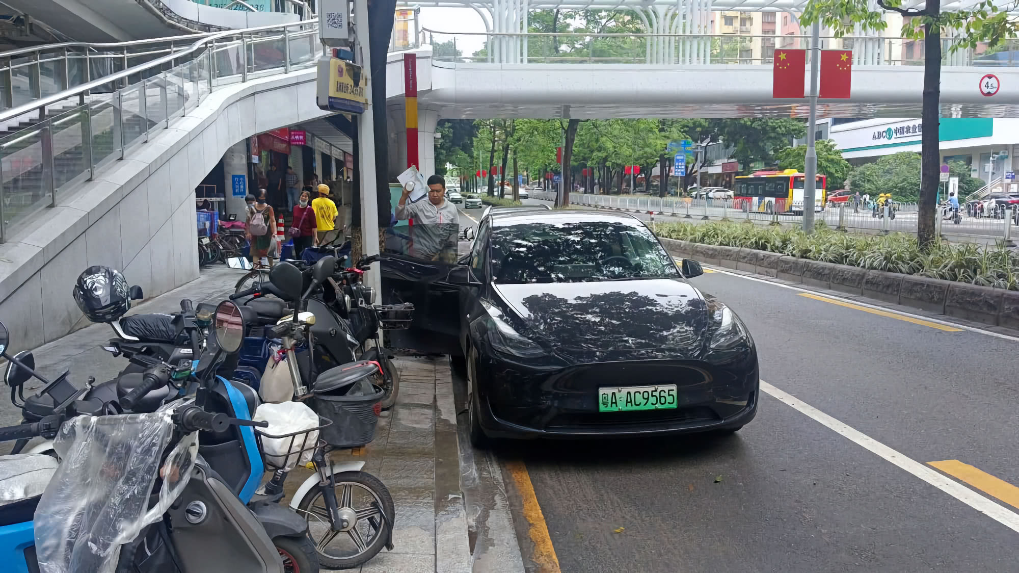 Чёрный электромобиль Tesla Model Y с зелёным номером A AC9565 припаркован у тротуара под пешеходным мостом; рядом стоят электросамокаты и мотоциклы, люди проходят мимо.