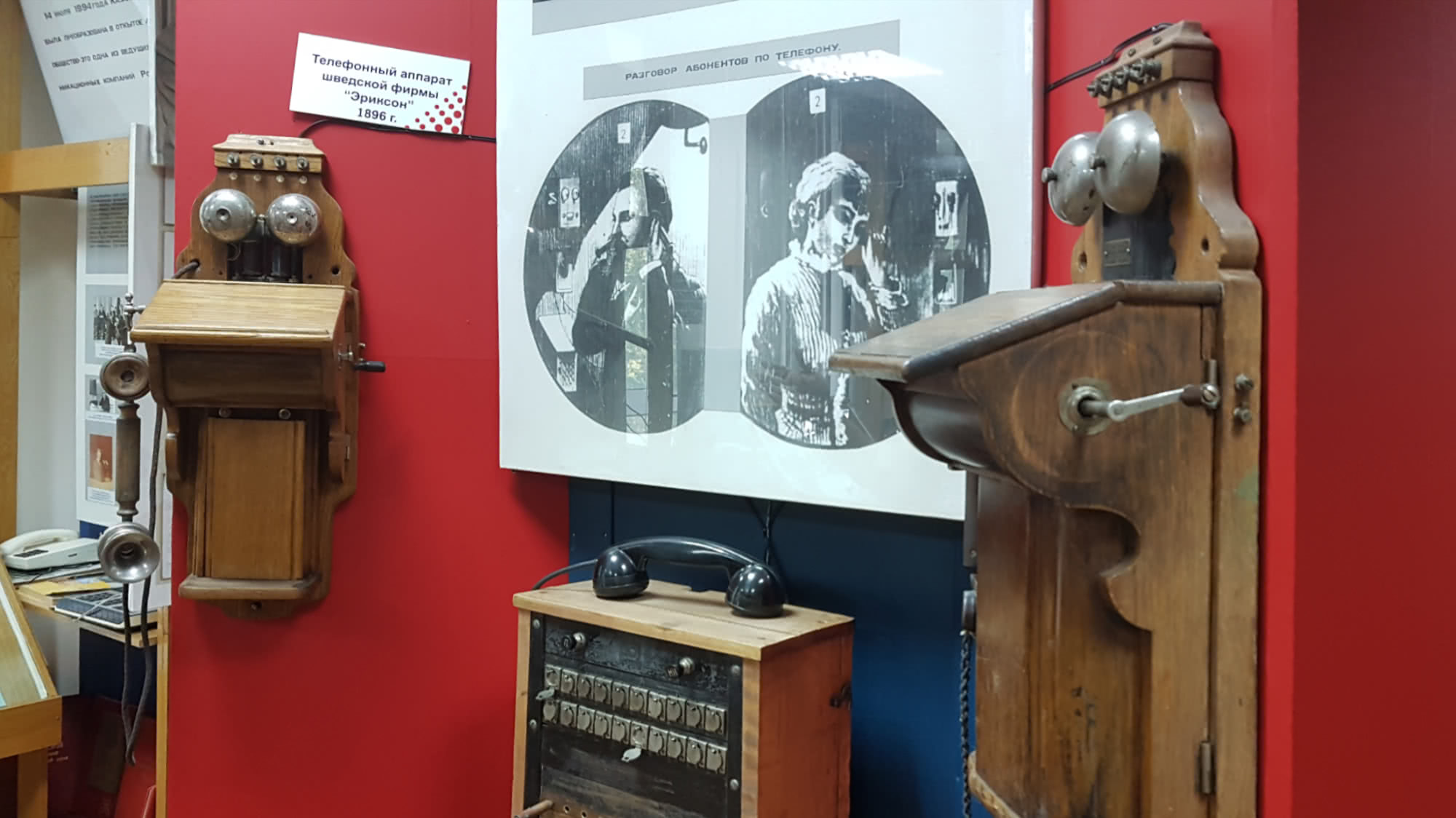 Музейные экспонаты: два деревянных настенных телефона 1896 г. фирмы «Эриксон» и телефонный коммутатор на красной стене под плакатом «Разговор абонентов по телефону».