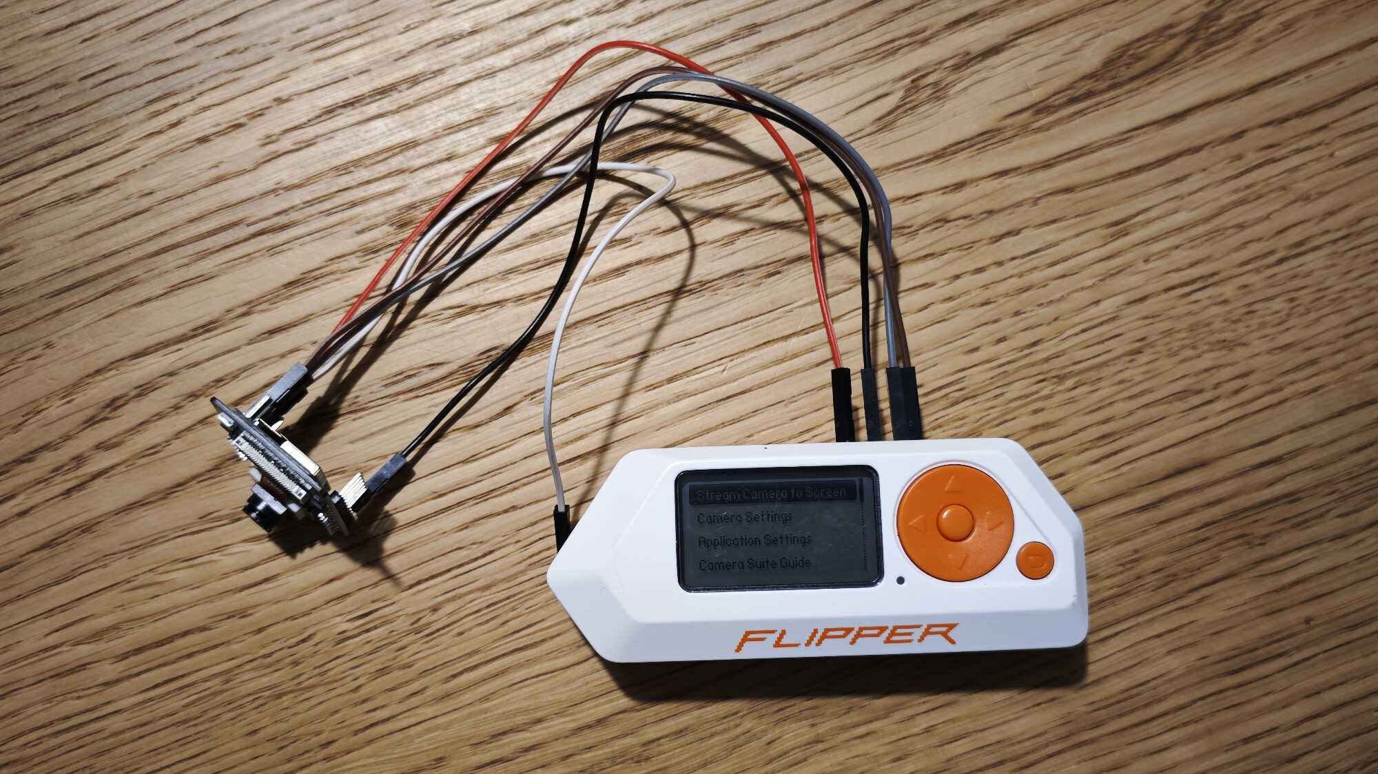Белый многофункциональный инструмент Flipper Zero с оранжевым джойстиком и экраном, на котором видно меню «Camera Settings», подключенный к плате через провода.