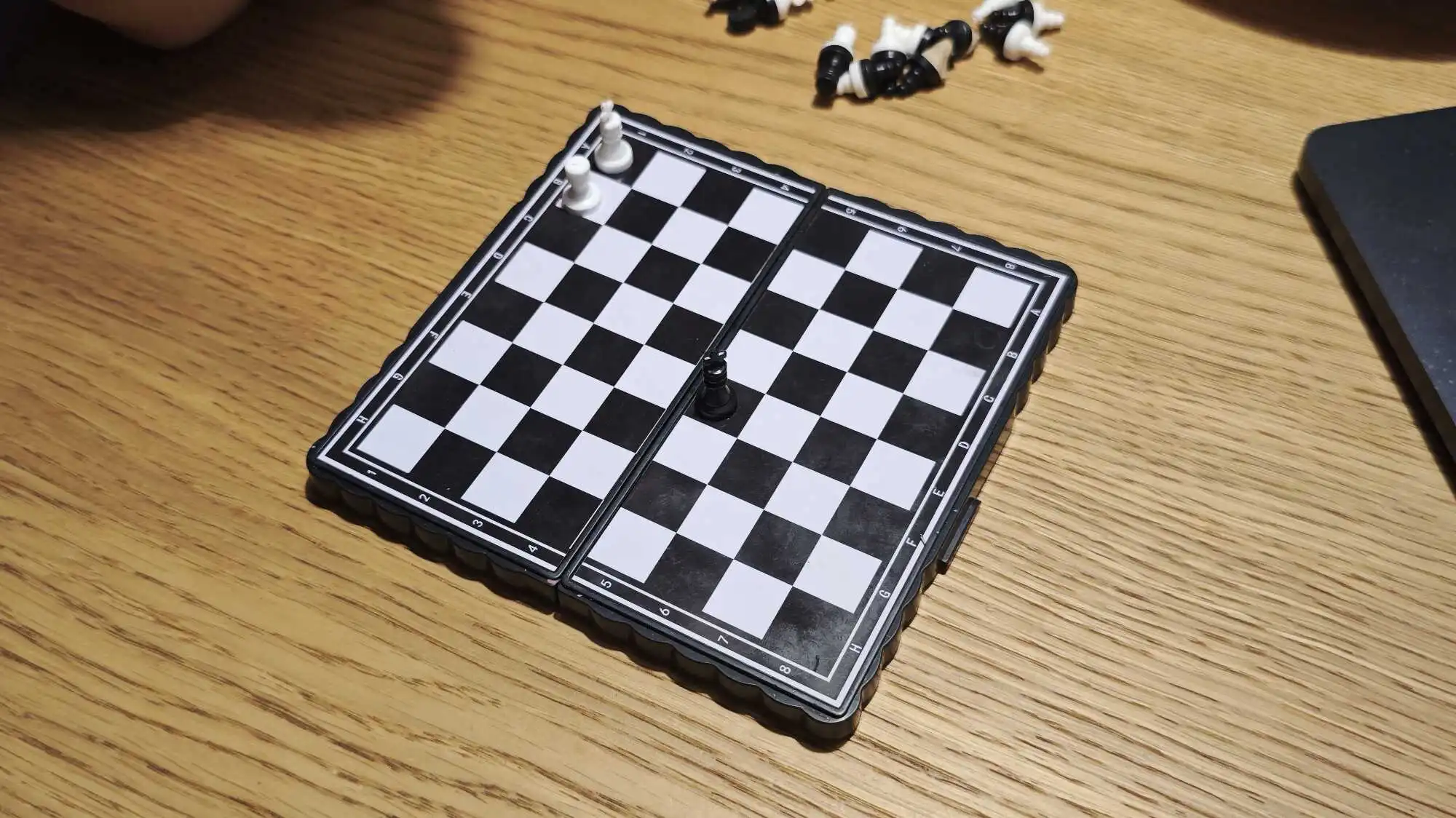 Складная шахматная доска с белыми и чёрными фигурами на деревянном столе; часть фигур разбросана рядом.