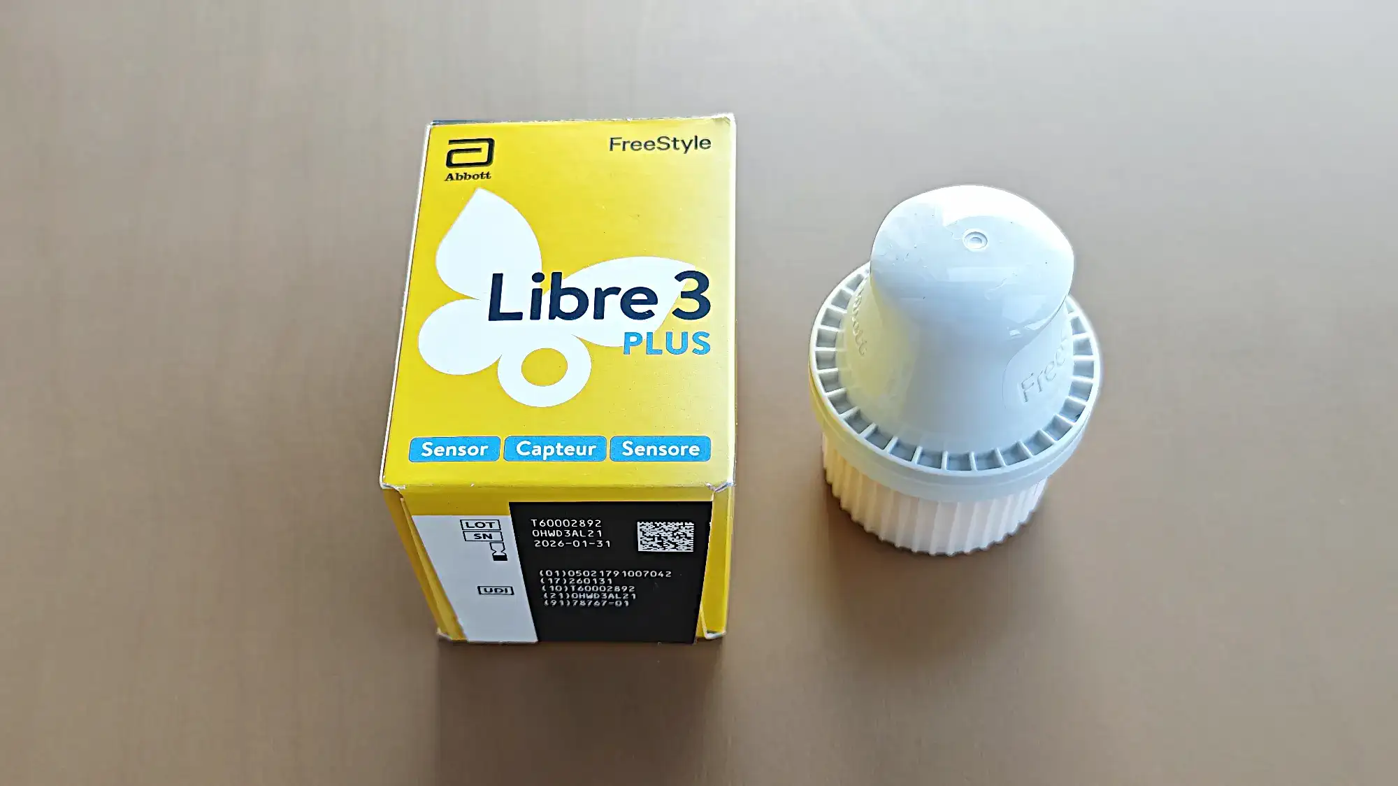 Желтая коробка FreeStyle Libre 3 Plus от Abbott с датчиком и белым пластиковым устройством рядом на светлом столе.