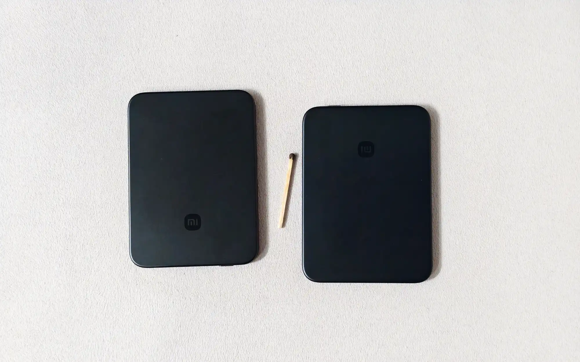 Два чёрных внешних аккумулятора Xiaomi с логотипом «mi» лежат на белой поверхности, между ними — спичка для сравнения размеров.