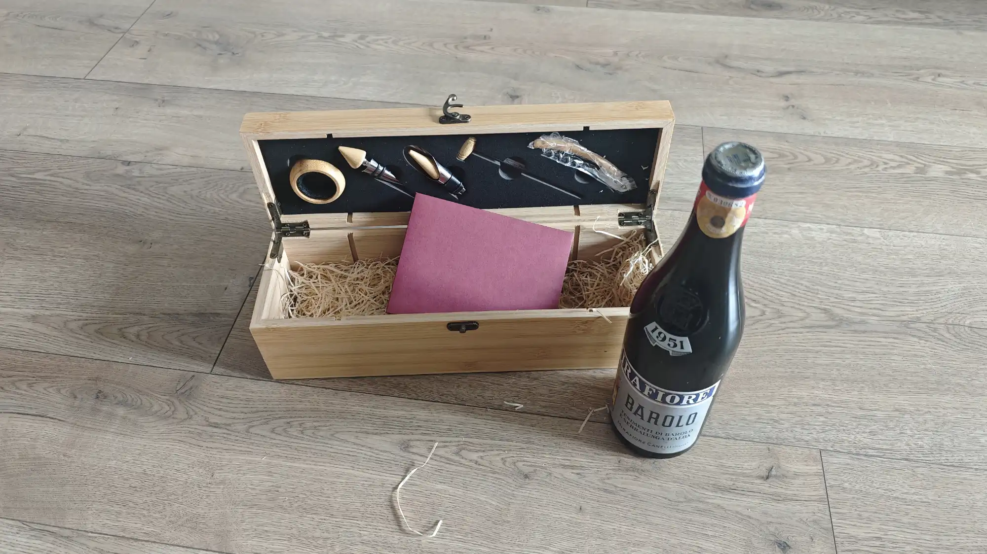 Открытая деревянная коробка с набором винных аксессуаров и бутылкой вина Barolo 1951 года на светлом деревянном полу.