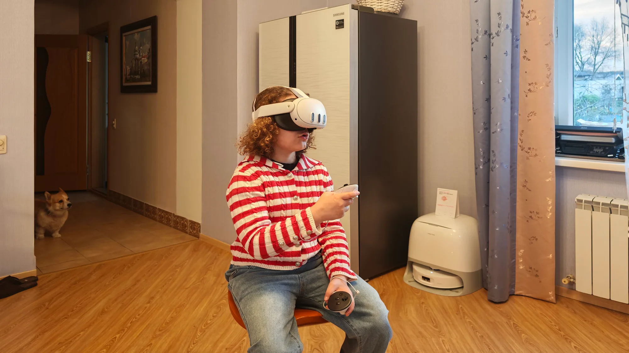 Девушка в красно-белой кофте сидит на стуле в комнате и играет в VR-игры, держа контроллеры.